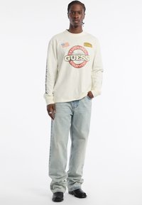 Langermige, off-white t-shirt met het "GUESS" logo in het rood en zwart, gecombineerd met lichtblauwe jeans. Model draagt zwarte schoenen en toont een ring.