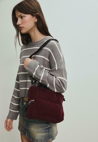Burgunderfarvet kordfløjspose med en rektangulær form og struktureret overflade, med en sort skulderrem og lynlås lukning. Båret med en gråstribet sweater.