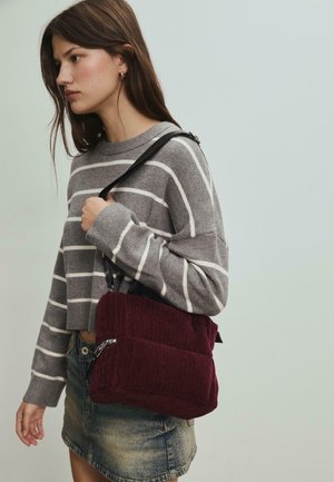 Burgunderfarbene Cordtasche mit rechteckiger Form und strukturierter Oberfläche, ausgestattet mit einem schwarzen Schulterriemen und einem Reißverschluss. Getragen zu einem grau gestreiften Pullover.