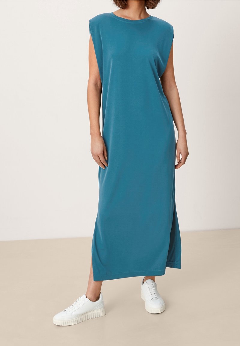 Femme portant une robe maxi sans manches bleu sarcelle avec des fentes latérales et des baskets blanches, debout devant un fond neutre.