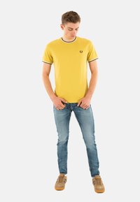 Camiseta de algodón amarilla con ribetes en blanco y negro, logo en el pecho izquierdo, combinada con jeans de color azul claro y zapatillas marrones.