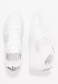 Zapatillas blancas con lados texturizados que presentan el nombre de la marca en un patrón repetido, cordones planos y un acento metálico en el talón.