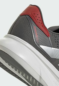 Zapatilla deportiva gris con parte superior de tela texturizada, acentos rojos, un logo plateado reflectante y una gruesa suela de goma blanca con un patrón de tracción.