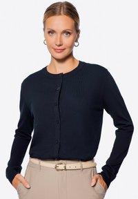 Modrá dlouhá svetr s dlouhým rukávem, kulatým výstřihem, předním zapínáním na knoflíky a ribbovanou strukturou, doplněná o béžové kalhoty s vysokým pasem a opaskem.
