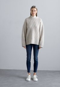 Yksinkertainen ja tilava beige neulepaita, jossa on ribbitekstuuri, yhdistettynä tummansinisiin slim-fit farkkuihin ja valkoisiin lenkkareihin. Yksinkertainen muotoilu, neutraali väriharmonia.