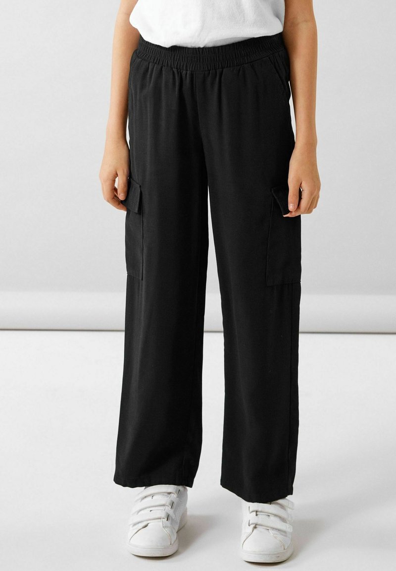 Enfant portant un pantalon cargo noir à jambes larges et des baskets blanches, debout devant un fond clair uni.