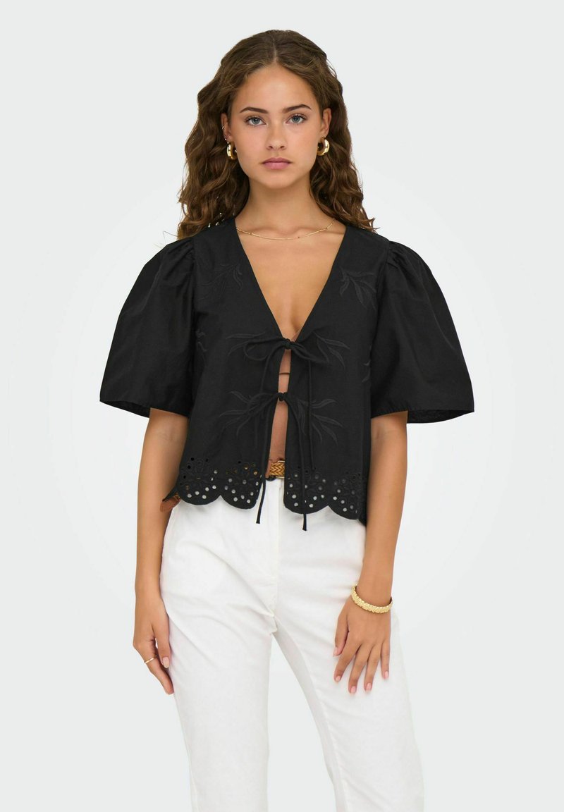 Schwarze Bluse mit kurzen Ärmeln, tiefem V-Ausschnitt, vorderer Bindung, gewelltem Saum, floralem Stickmuster und Spitzenverzierungen. Kombiniert mit weißen Hosen.
