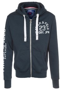 Sudadera con cremallera en azul marino, con cordones blancos, bolsillos frontales y texto blanco "TRACK & FIELD 23 SUPERDRY. JPN" en el pecho y la manga.