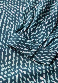 Tessuto verde a motivi con un design a onde. Presenta un polsino con bottoni e arricciature e una texture liscia. Contrasto di colore tra tonalità chiare e scure.