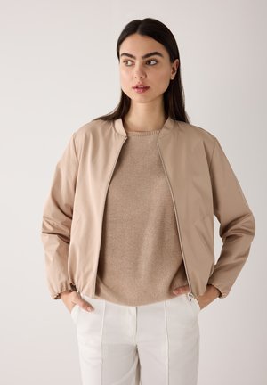 Vrouw met lang donker haar draagt een beige jas met rits over een gebreide trui en witte broek, staat met haar handen in de zakken en kijkt opzij.