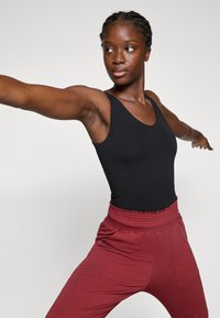 Γυναίκα με μαύρο τοπ και κόκκινο παντελόνι, τεντώνει τα χέρια πλατιά, κοιτάζοντας προς το πλάι, με φόντο απλό ανοιχτό.