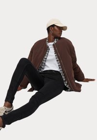 Chaqueta bomber acolchada marrón sobre una camisa de cuadros, combinada con vaqueros ajustados negros y zapatillas ligeras. Una gorra y pendientes de aro completan el look.