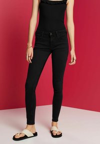 Jeans ajustados negros con cintura alta y detalles de costura sutil, combinados con sandalias de banda cruzada color crema sobre un fondo rosa.