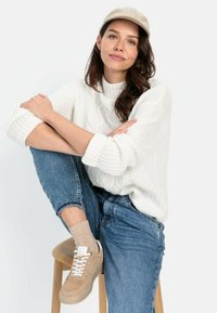 Weißer Strickpullover mit strukturiertem Muster, blaue Jeans, beige Sneakers und eine hellfarbige Kappe. Das Model sitzt auf einem Holzstuhl.