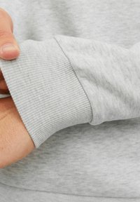 Manga de sudadera gris claro con puño acanalado, que muestra una textura suave y un sutil patrón jaspeado. Mano parcialmente visible.