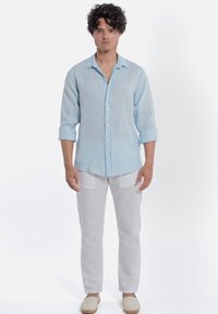 Camicia di lino azzurro chiaro con maniche arrotolate, bottoni sul davanti, abbinata a pantaloni di lino bianchi e scarpe espadrillas beige.
