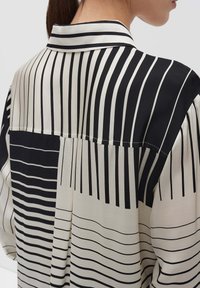 Schwarz-weiß gestreiftes Hemd mit variierenden Streifenbreiten und einem Kragen, hergestellt aus leichtem Stoff mit glatter Textur.