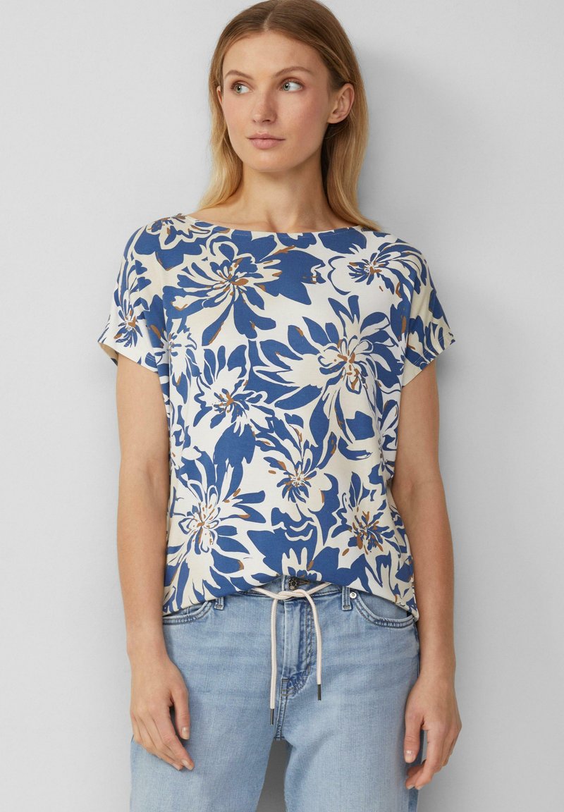 Haut en imprimé floral crème avec des accents bleus et dorés, dotée de manches courtes, d'un col rond et d'une coupe décontractée. Associé à un jean en denim clair.