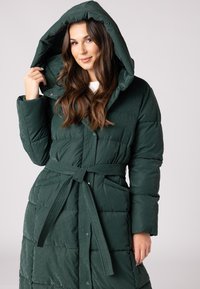 Groene pufferjas met capuchon, gewatteerd ontwerp, tailleband en zijzakken, met een glanzende textuur en knoopsluitingen.
