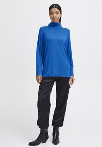 Pulover albastru cu guler înalt, fabricat dintr-un material moale, cu o croială supradimensionată, manșete ribbate. Pantaloni cargo negri, cu buzunare laterale și o textură fină, asortați cu cizme până la gleznă.