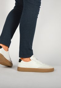 Des baskets en cuir blanc avec une semelle en caoutchouc et un accent noir au talon, présentant un design à lacets et un branding subtil sur le côté.