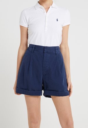 Femme portant un polo blanc à manches courtes avec logo bleu et un short bleu marine taille haute avec revers, sur fond uni.