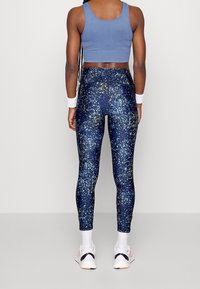 Vrouw in een blauwe crop top en hoge navy leggings met een gele en lichtblauwe spikkelpunt, witte sneakers en polsbandjes, gezien van achteren.