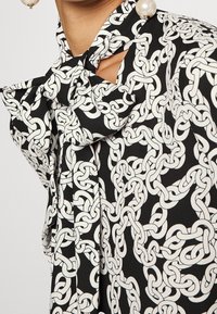 Blusa negra con un patrón de cadenas blancas, que cuenta con un cuello atado y un detalle de hombro descubierto. Tela suave con una silueta fluida.