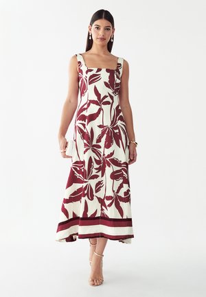 Mujer con un vestido blanco sin mangas con grandes patrones de hojas rojas, sosteniendo un bolso blanco de mano y con tacones beige con tiras en el tobillo.