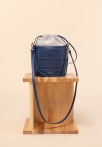 Borsa a secchiello in pelle blu con una parte superiore in cordino grigio, accessori dorati, tasca frontale e tracolla regolabile. Posizionata su un supporto di legno.