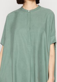 Zelená blůza z lehkého materiálu, má kulatý výstřih a krátké rukávy, s částečným knoflíkovým zapínáním a volným střihem.