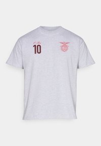 Γκρι βαμβακερό T-shirt με κόκκινο κεντητό κείμενο "Ο Ρέι" και τον αριθμό "10", καθώς και ένα κόκκινο έμβλημα στην αριστερή περιοχή του στήθους.