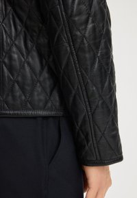 Primo piano di una manica e di una cucitura laterale di una giacca di pelle nera trapuntata su tessuto scuro, con una mano parzialmente visibile in basso.