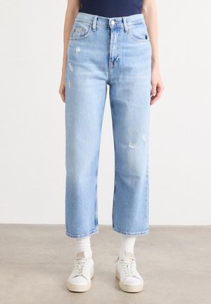 Persoon draagt lichtblauwe, rechte, high-waist jeans met kleine versleten details, witte sokken en witte sneakers, staand op een grijze vloer.