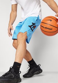 Svetlo modri košarkarski šortji z črnim logotipom, strukturirano blago in stranskimi razporki. V kombinaciji s črnimi športnimi čevlji in nogavicami.
