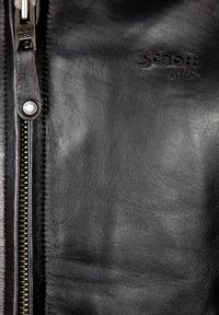 Schwarze Lederjacke mit glatter Textur, ausgestattet mit einem silbernen Reißverschluss und einem geprägten Logo mit der Aufschrift "Schott." Nahaufnahme der Materialien.