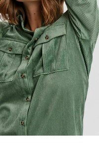 Camicia button-up in corduroy verde con una finitura testurizzata, dotata di due tasche sul petto, bottoni marroni e colletto.