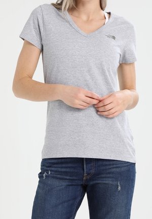 Person trägt ein hellgraues V-Ausschnitt T-Shirt mit kurzen Ärmeln und dunkelblaue Jeans mit leichter Abnutzung, steht mit locker verschränkten Händen.