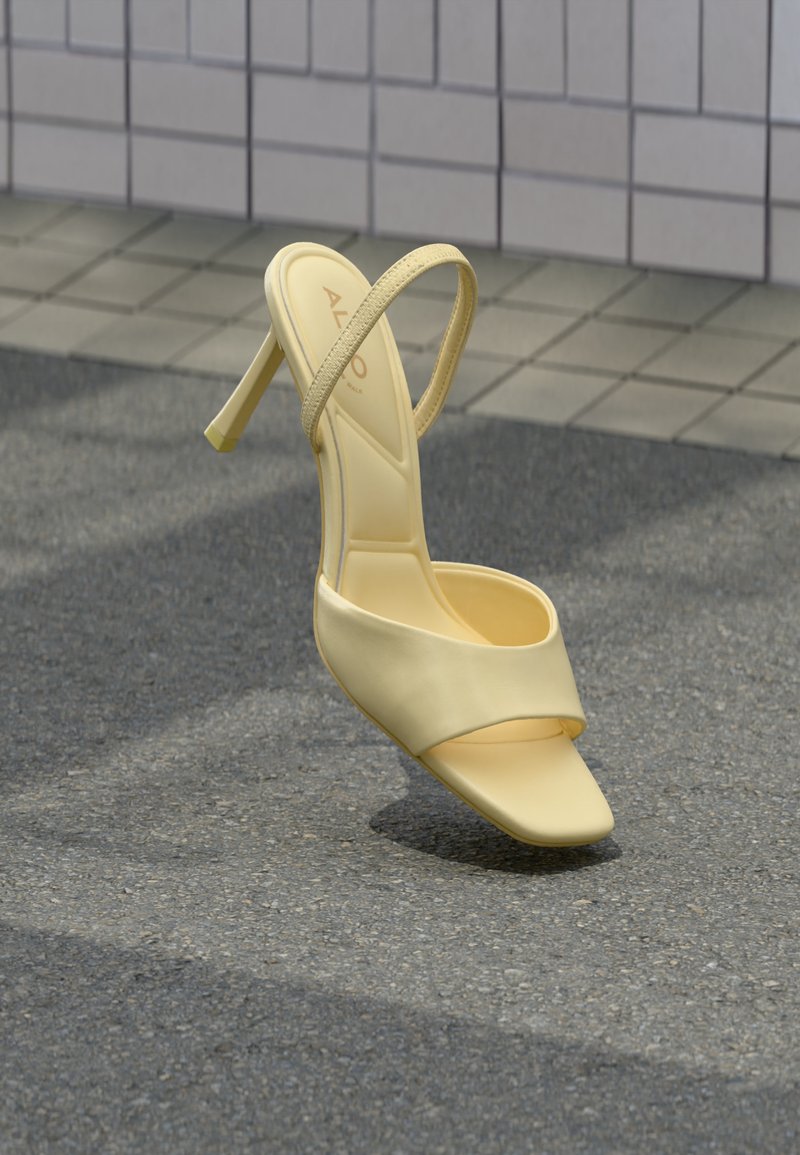Sandale à talon haut jaune avec une fine bride et un bout ouvert, positionnée verticalement sur un pavement texturé près d'un mur carrelé.