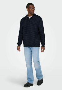 Dunkelblauer Kabelstrickpullover mit Kragen, kombiniert mit hellblauen Jeans und schwarzen Schuhen. Der Pullover hat gerippte Bündchen und einen gerippten Saum.