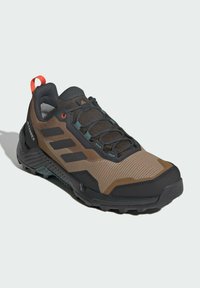 Brown and black trail running shoe with textured upper, reinforced toe, flexible sole, and contrasting orange accents. Features lacing system.