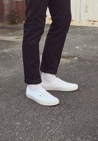 Weiße Leinensneaker mit Schnürverschluss, Gummisohlen und dezentem Seitenlogo, kombiniert mit schwarzen Hosen und weißen Knöchelsocken.