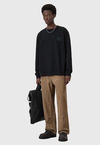 Sudadera negra con logo redondo, combinada con pantalones de pana color beige con corte ancho y bolsillos laterales. Bolsa negra y zapatos oscuros.