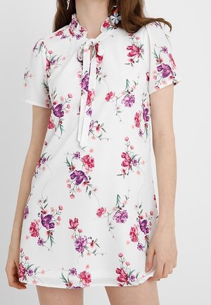 Robe blanche à manches courtes avec un motif floral en rose et violet, dotée d'un col mandarin et d'un détail noué au niveau de l'encolure.