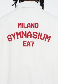 Balta striukė su raudonais siuvinėjimais: "MILANO GYMNASIUM EA7" ant nugaros, su klasikinio stiliaus apykakle ir glotnia tekstūra.