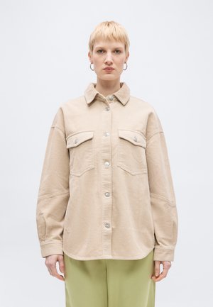 Lyshåret person med kort hår iført beige fløjlsjakke med lommer med klap og lysegrønne bukser, stående mod en ensfarvet baggrund.