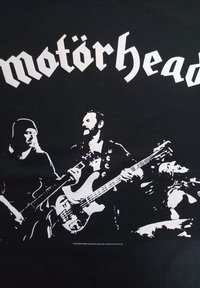 Camiseta negra con una ilustración gráfica blanca de tres músicos con instrumentos. El nombre de la banda "Motörhead" está exhibido de manera prominente en la parte superior.