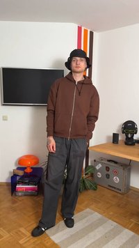 Barna cipzáros kapucnis pulóver, szürke, bő szabású nadrág és fekete kalap. Figyelemre méltó narancssárga kiegészítő asztal és csíkozott fal a háttérben. Minimalista dekoráció.