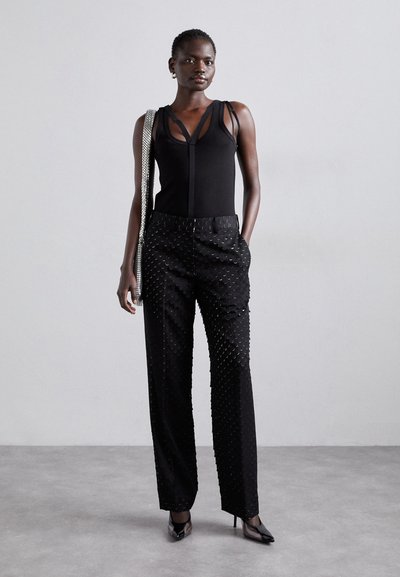 Fekete, ujjatlan felső, egyedi pánttervezéssel, struktúrált fekete nadrággal, amelyen kör alakú minták találhatók. A modell egy metálos kézitáskát tart.