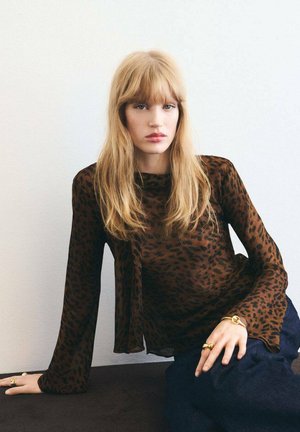 Blusa trasparente marrone con stampa leopardata nera, caratterizzata da maniche lunghe e un design fluido. Indossata con jeans di denim e dettagli in gioielli dorati.